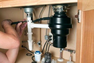Plumber repairing a garbage disposal beneath a kitchen sink in Thornton